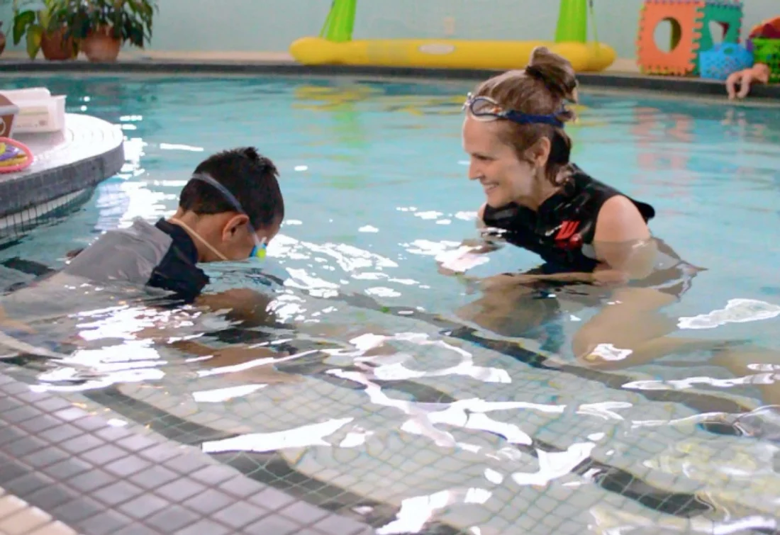 Tessa Rhodes smiling and encouraging a young boy as he puts his face in the water